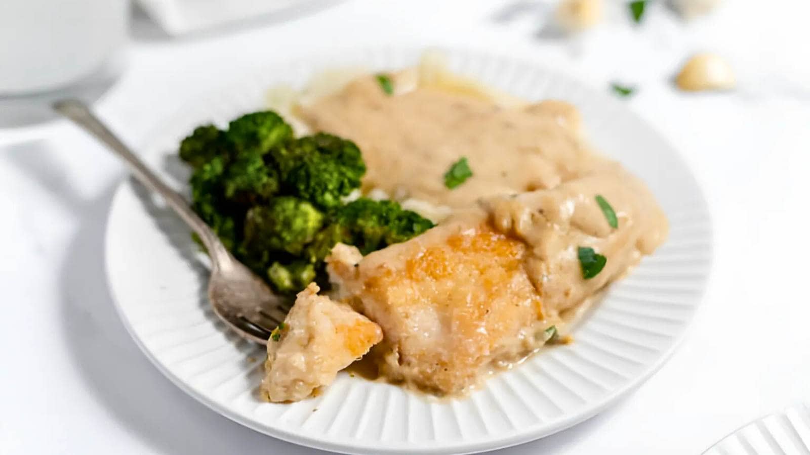 A plate with fish fillet covered in creamy sauce, a side of steamed broccoli, and a fork, on a white dish.