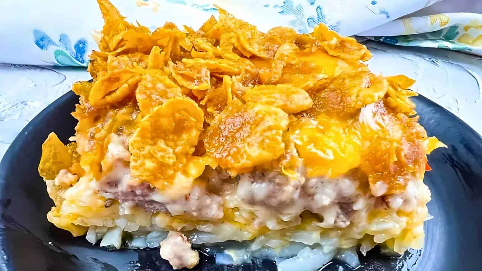 A close-up of a layered casserole on a black plate. It features ground meat, potatoes, cheese, and a topping of cornflakes. A patterned cloth is in the background.