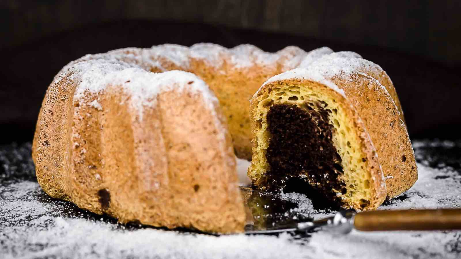 A bundt cake with a slice cut, showing a chocolate center. The cake is sprinkled with powdered sugar and a cake server is partially visible.