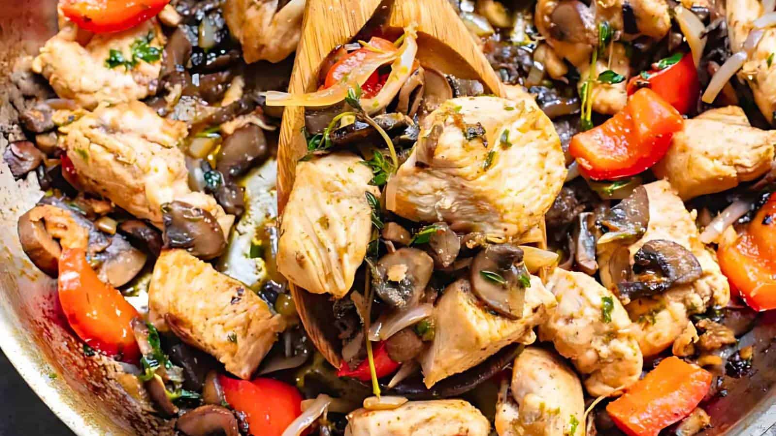A close-up of a pan containing cooked chicken pieces, mushrooms, red bell peppers, and herbs, being stirred with wooden utensils.