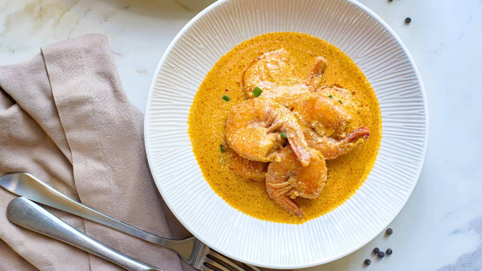 A white bowl of creamy orange shrimp curry garnished with herbs, beside a folded napkin with a fork and spoon on a marble surface.