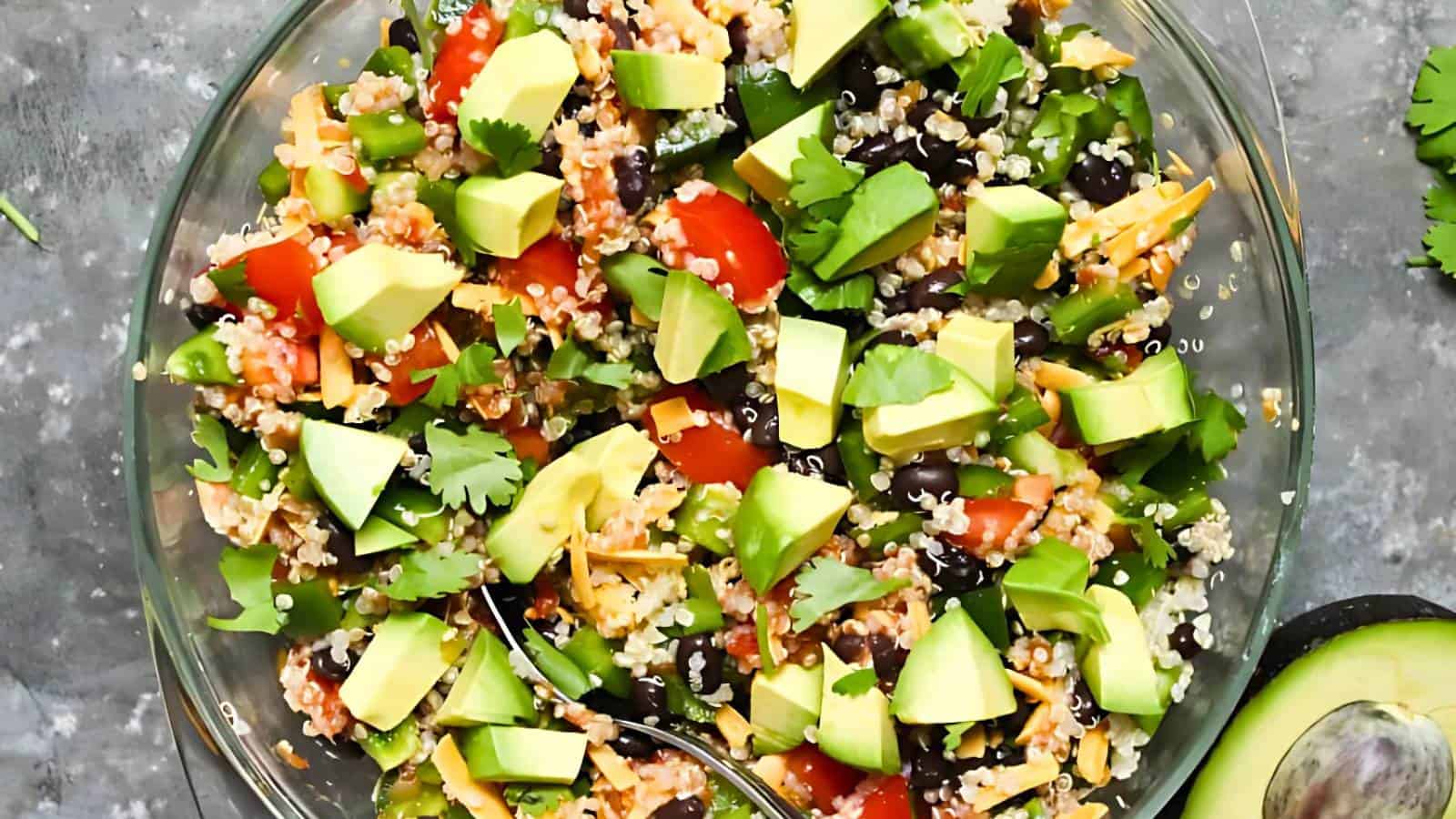 A glass bowl filled with quinoa salad, featuring diced avocado, tomatoes, black beans, quinoa, cheese, and cilantro, with a spoon beside it. A halved avocado is nearby.