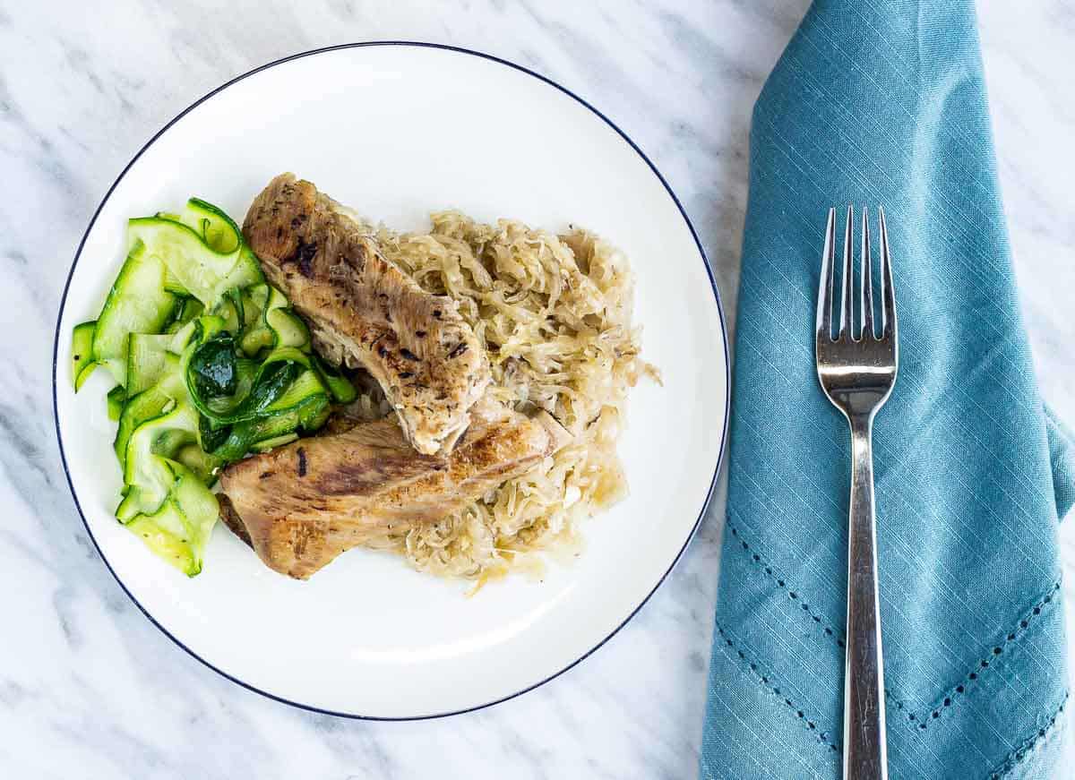 Plate with ribs, sauerkraut, and sliced zucchini next to a blue napkin and fork on a marble surface.