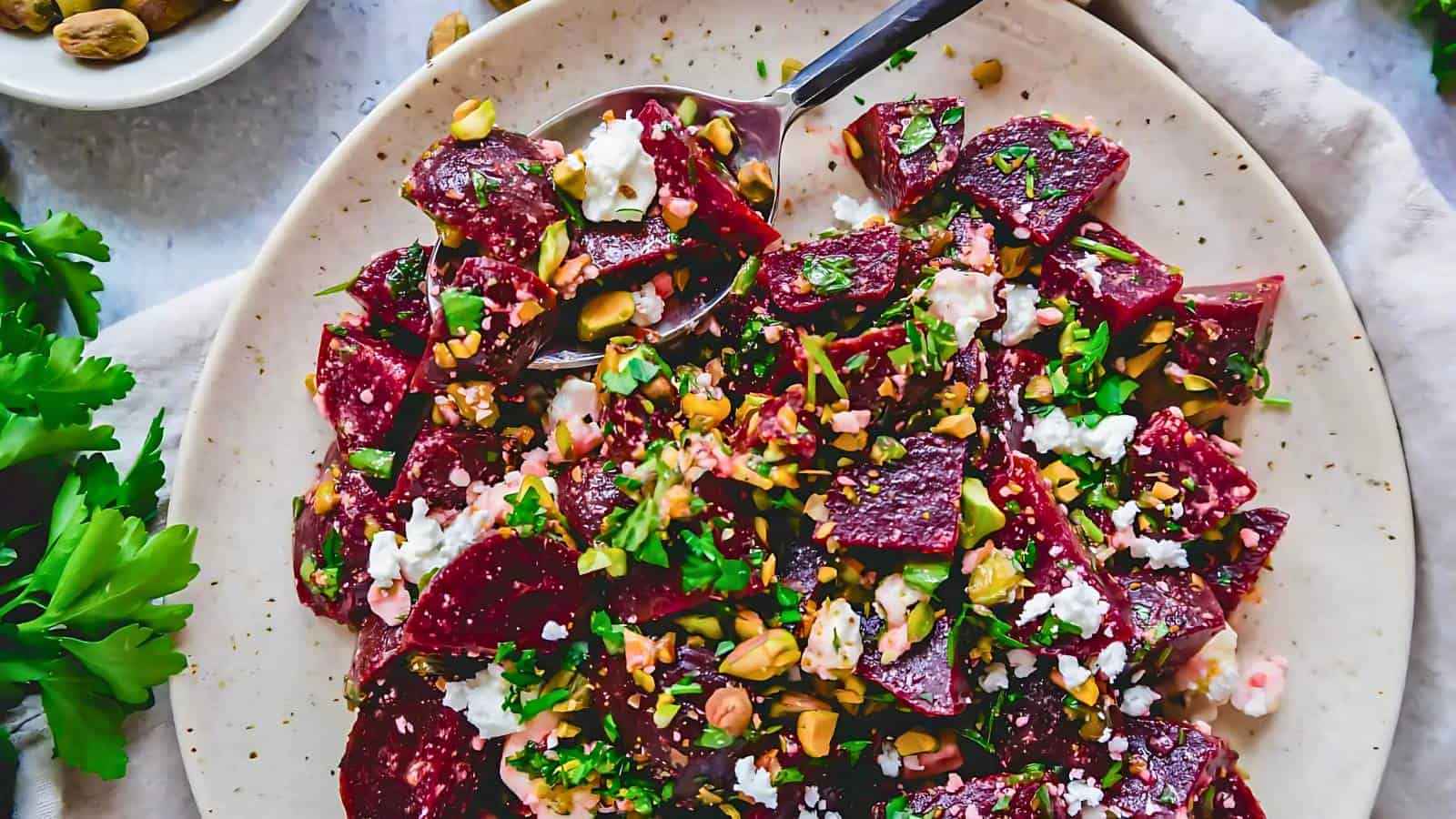 Plate of beet salad with crumbled cheese, chopped nuts, and herbs, accompanied by a serving spoon.