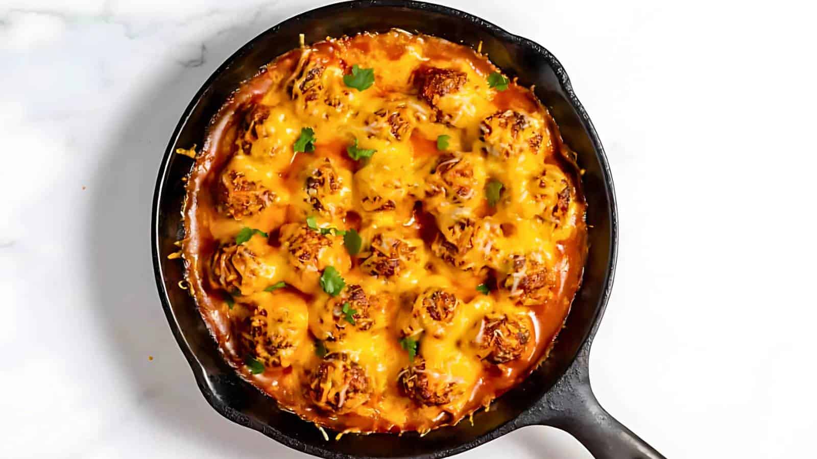 A cast iron skillet filled with meatballs covered in melted cheese and garnished with herbs, set on a white marble surface.