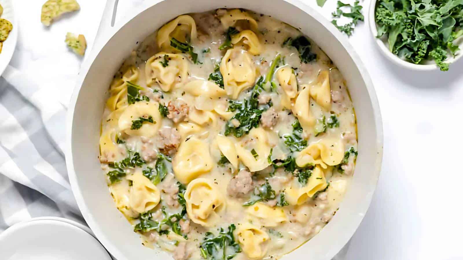 A pot of creamy tortellini with sausage and kale, placed on a white surface with a striped napkin and small bowls of tortilla chips and greens beside it.