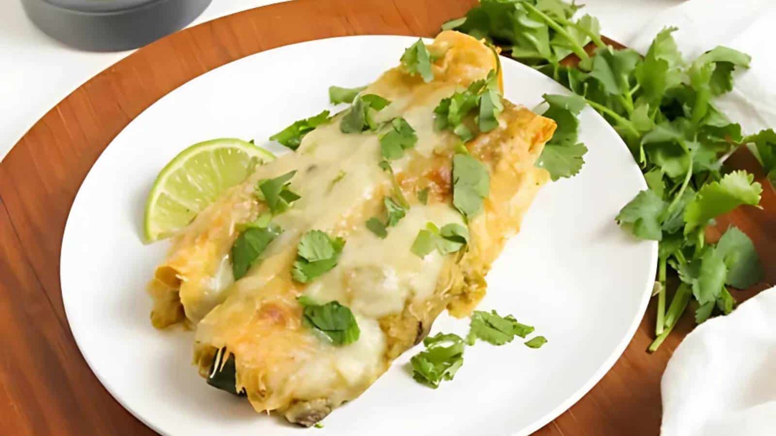 Cheesy enchiladas topped with cilantro on a white plate, accompanied by a lime slice. Parsley is placed beside the plate on a wooden surface.