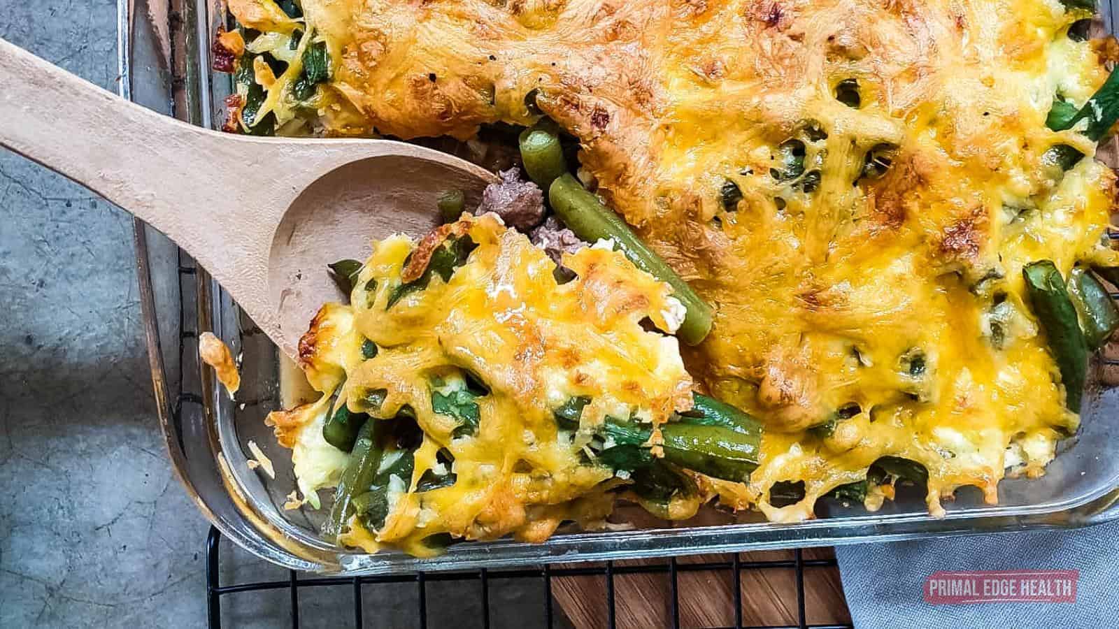 A baked casserole in a glass dish containing melted cheese, green beans, and spinach, with a wooden spoon serving a portion.