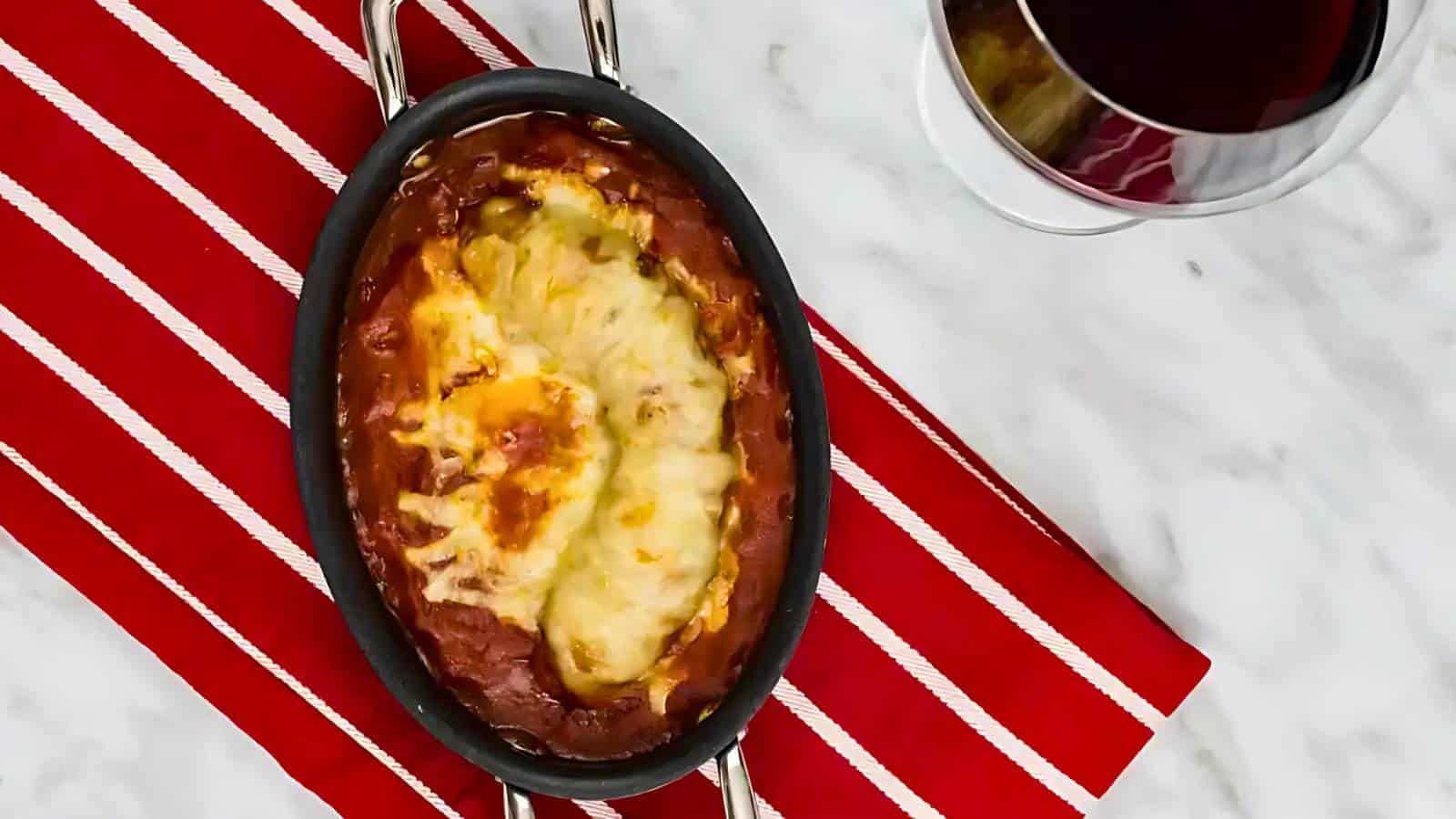 Baked dish with melted cheese on top, set on a red and white striped cloth next to a glass of red wine on a marble surface.