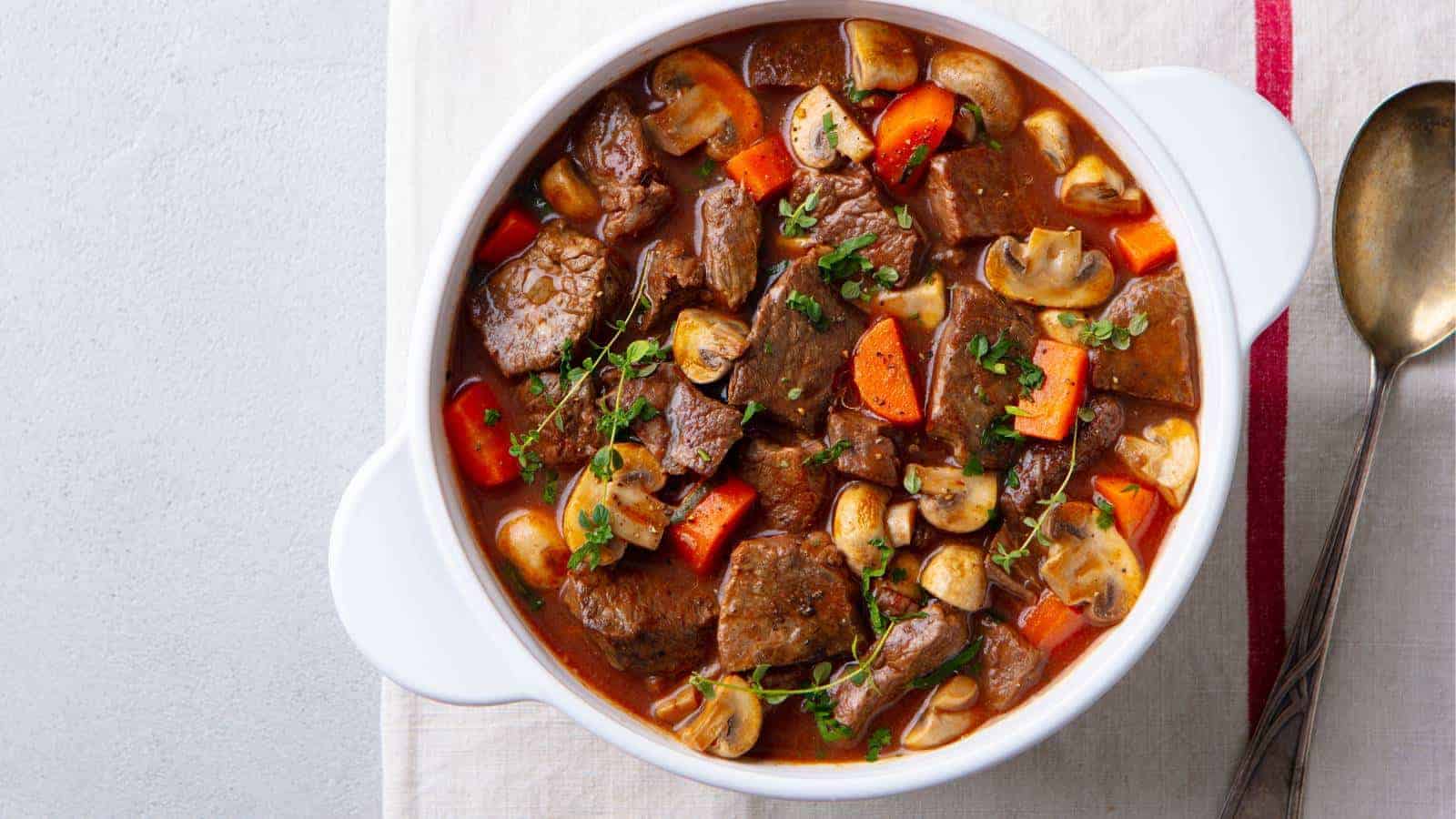 A bowl of beef stew with chunks of beef, mushrooms, and carrots in a rich brown sauce, garnished with herbs. A spoon is placed beside the bowl on a white surface.