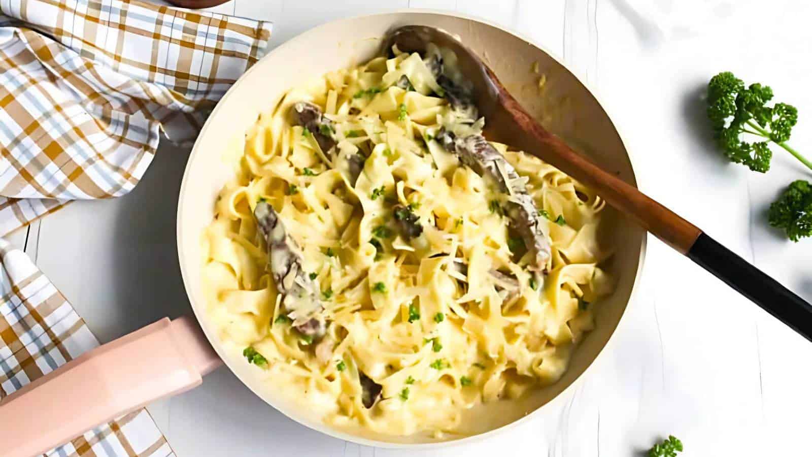 A pan of creamy pasta with slices of sausage, garnished with chopped herbs and grated cheese. A wooden spoon rests on the side. A checked cloth and parsley are nearby.