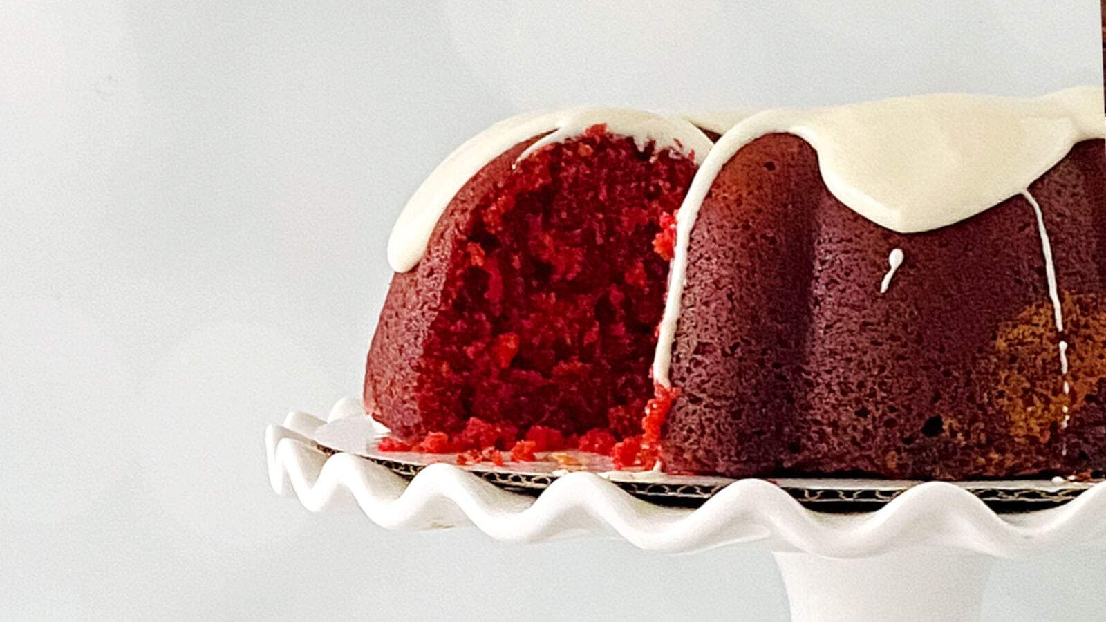 A red velvet bundt cake with white icing is displayed on a white fluted cake stand. A slice is missing, revealing the cake's red interior.
