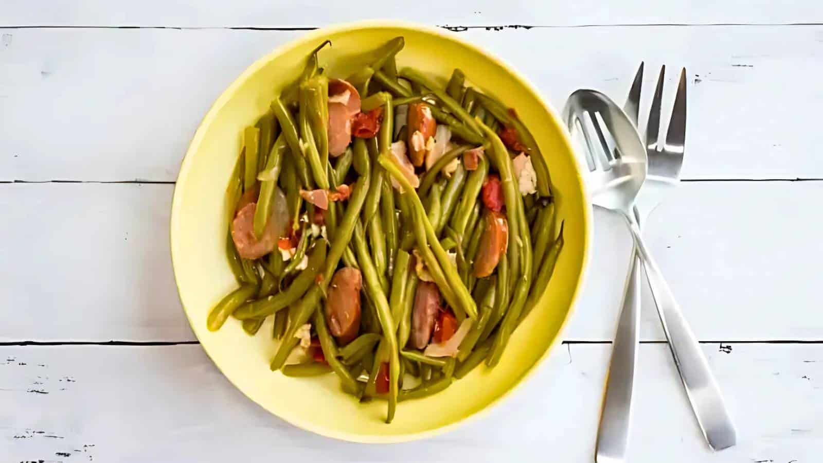 A yellow plate of cooked green beans mixed with sliced sausages and diced tomatoes. Two serving utensils are placed beside the plate on a white wooden surface.