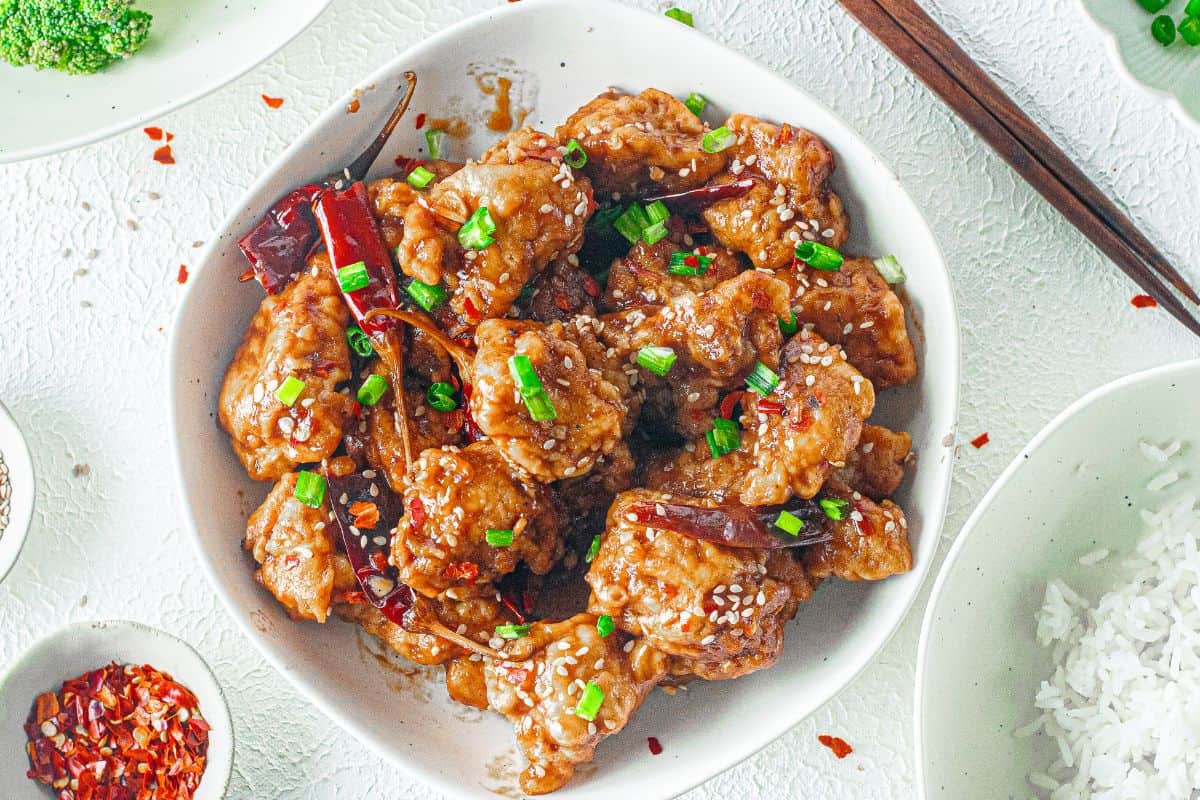 A bowl of crispy General Tso chicken pieces garnished with sesame seeds, chopped green onions, dried red chilies, and served with rice and broccoli on the side.