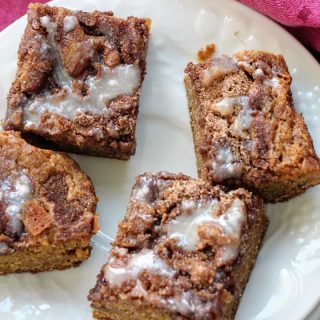 Four square pieces of glazed maple blondies on a white plate, placed on a wooden surface with a pink fabric nearby.