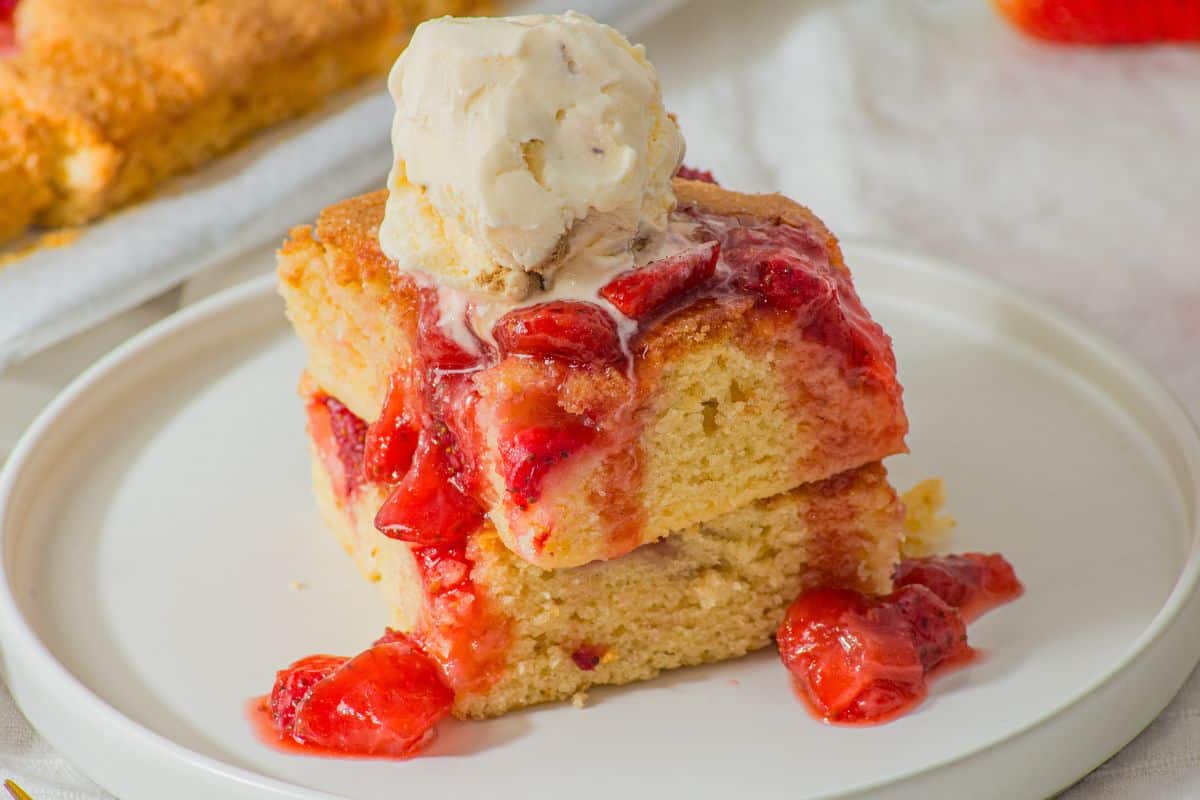 Two pieces of strawberry-topped cake stacked on a plate, with a scoop of vanilla ice cream on top, evoke memories of a favorite strawberry sheet cake recipe. More cake tantalizes in the background.