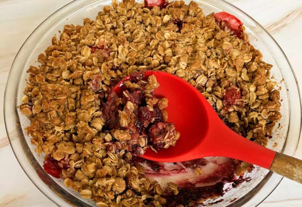 A round glass dish containing a berry crumble with oats, a portion scooped out, revealing the red berry filling underneath. A red spoon rests inside.