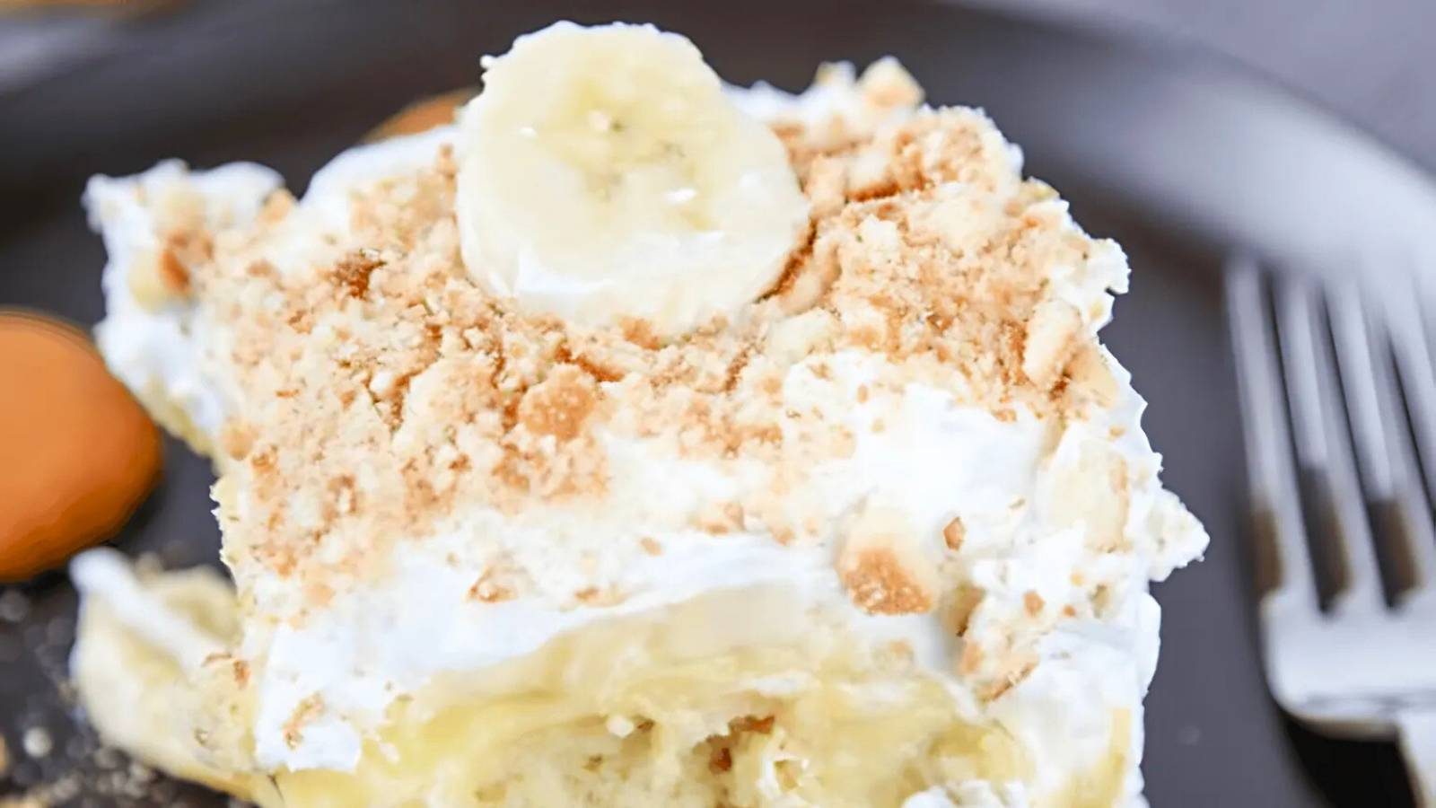 A slice of banana pudding dessert topped with whipped cream, crumbled cookies, and a banana slice on a dark plate beside a fork.