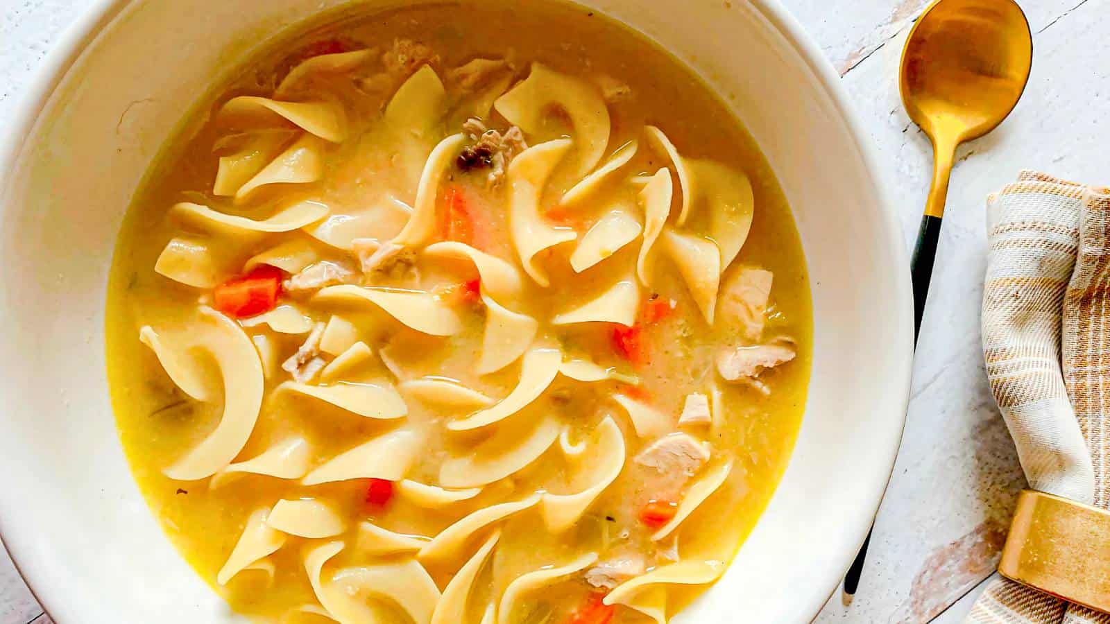 Bowl of chicken noodle soup with wide egg noodles, diced chicken, and carrot pieces. A spoon and striped napkin are placed beside the bowl on a wooden surface.