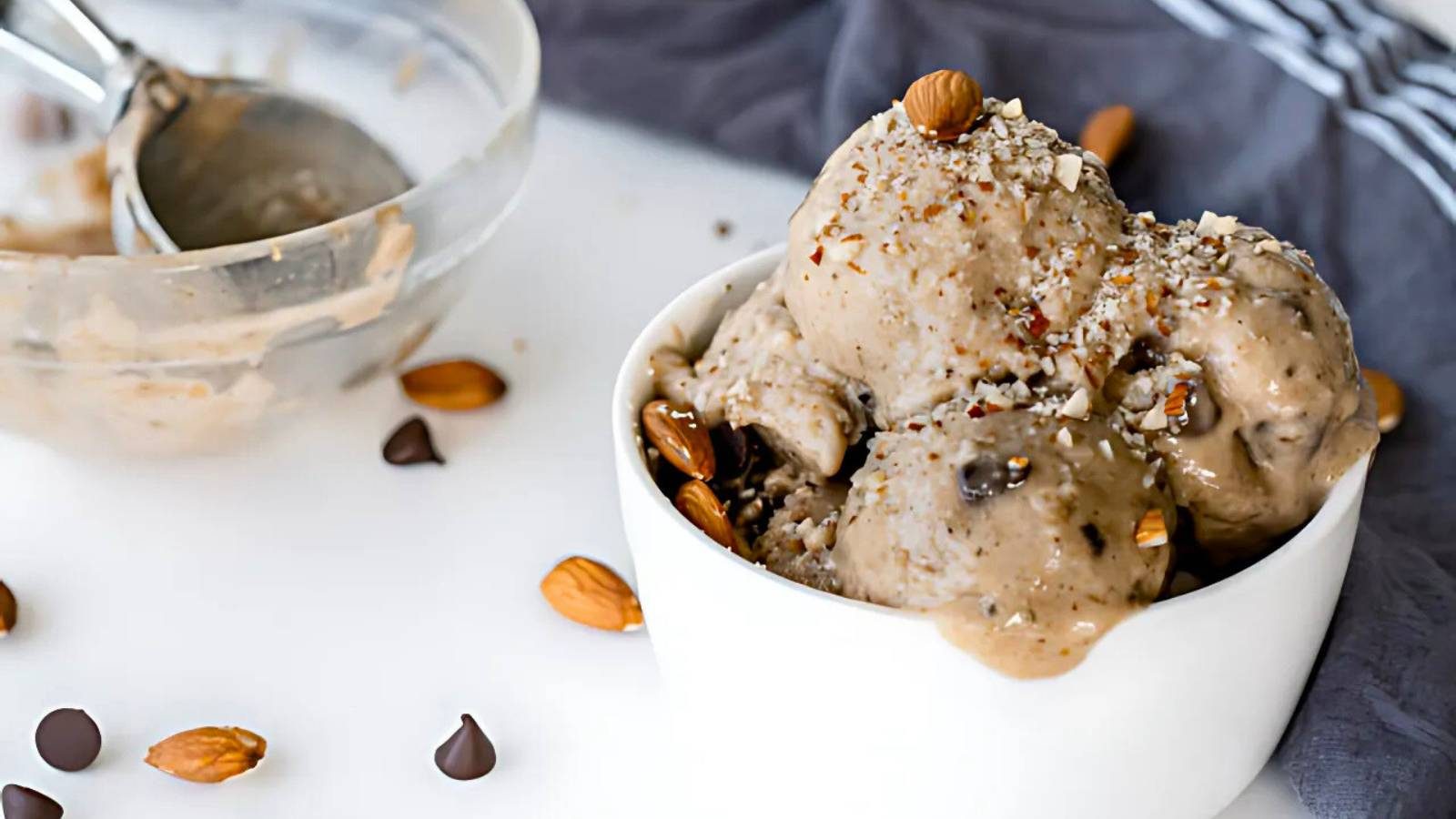 A bowl of ice cream with chocolate chips and chopped almonds on top, surrounded by scattered almonds and chocolate chips, with a glass bowl and ice cream scoop in the background.