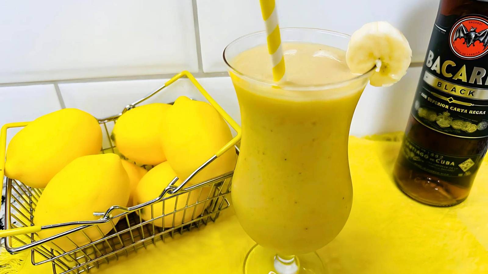 A glass of yellow smoothie with a banana slice and straw, beside a basket of lemons and a bottle of Bacardi Black rum on a yellow surface.