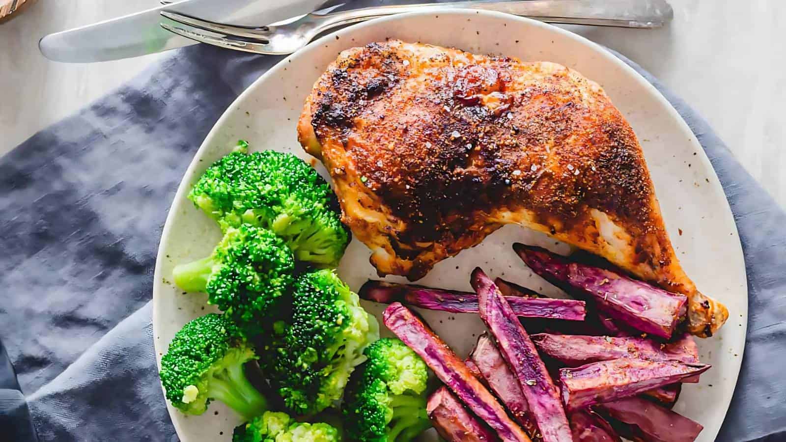 A plate with baked chicken leg, steamed broccoli, and purple sweet potato fries, with a fork and knife placed above the plate.