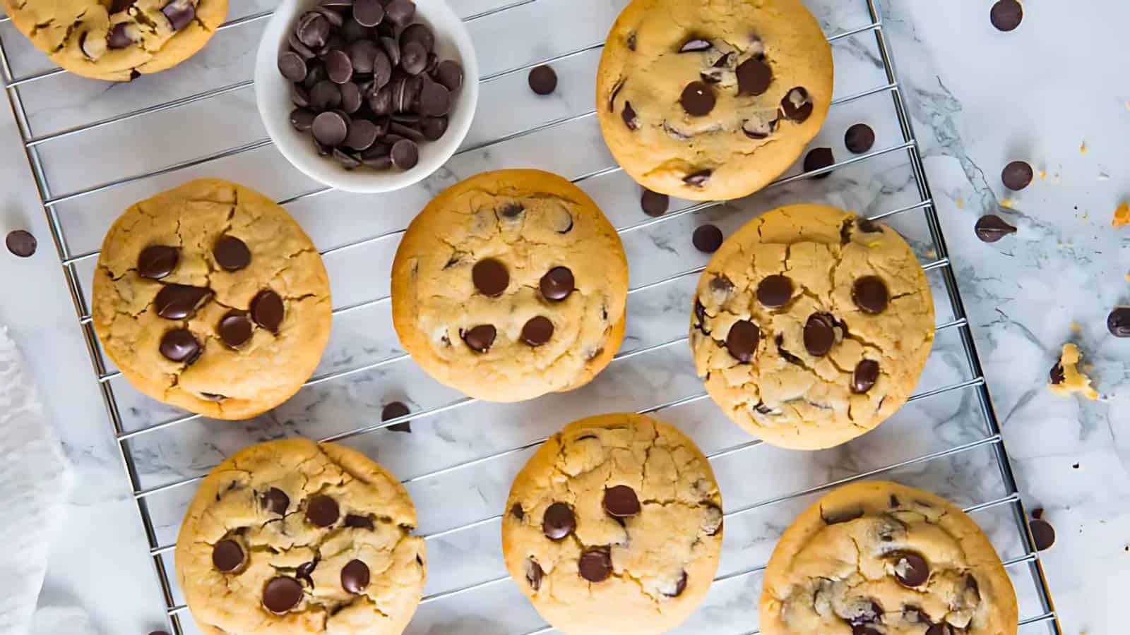Chocolate chip cookies on a cooling rack with a small bowl of chocolate chips, photographed from above on a marble surface.