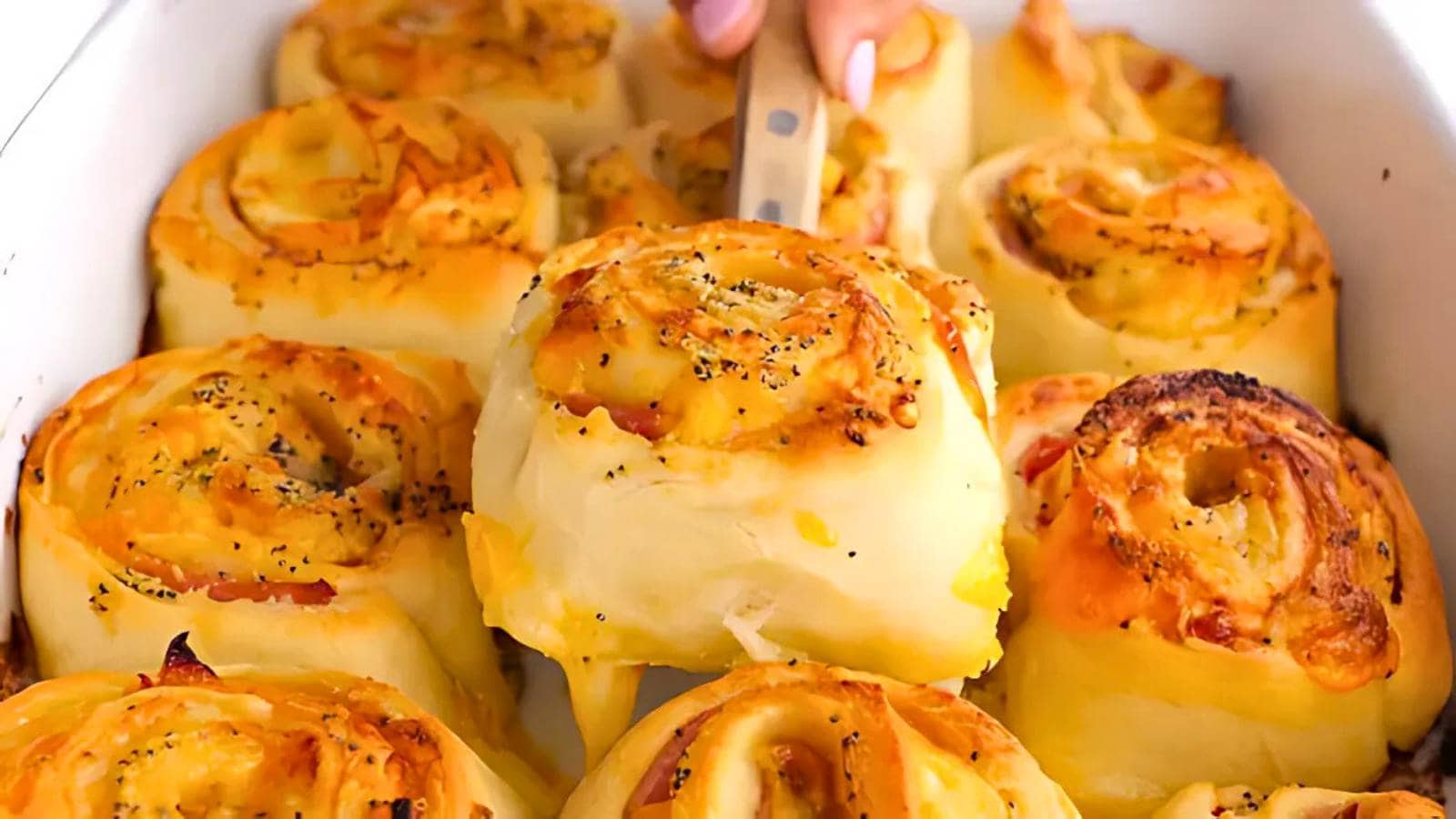 A baking dish filled with several savory cheese and ham roll-ups, with one piece being lifted out using a spatula.