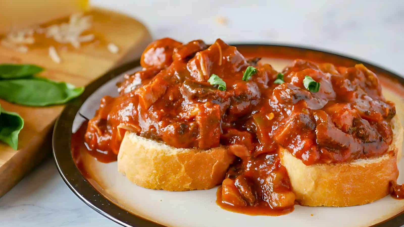 Two slices of bread topped with a thick, chunky tomato-based meat and mushroom stew, garnished with basil, served on a white plate.