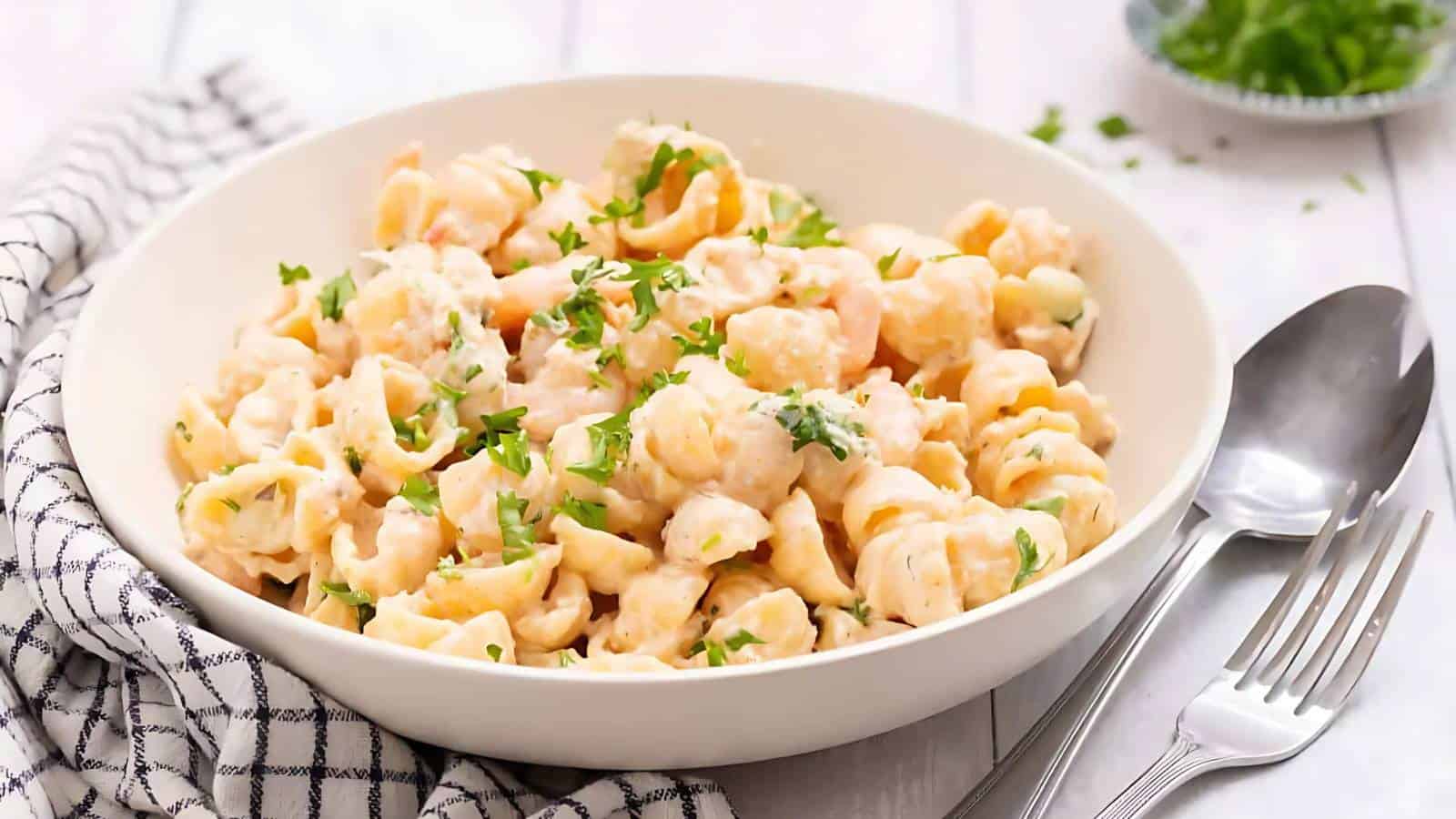 A white bowl filled with creamy shell pasta, garnished with chopped parsley, sits beside a fork, spoon, and checkered napkin on a light-colored table.