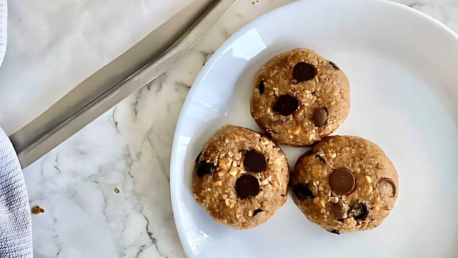 Three unbaked chocolate chip cookies on a white plate set on a marble countertop, with a knife and towel nearby.