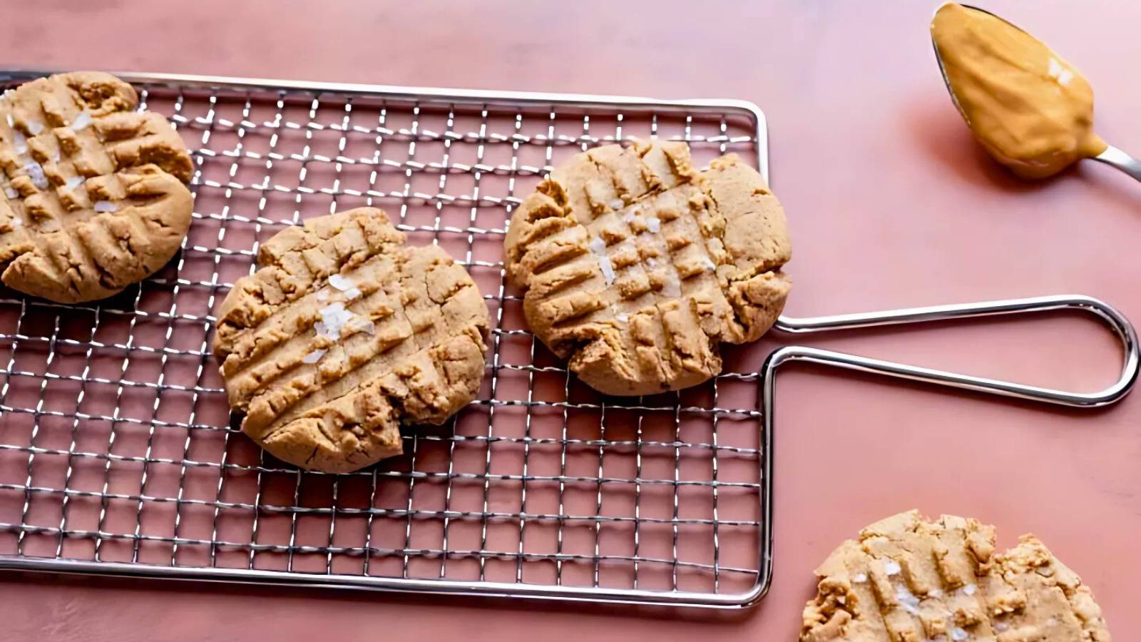 Four peanut butter cookies with crisscross fork marks are on a cooling rack, with a spoonful of peanut butter nearby on a pink surface.