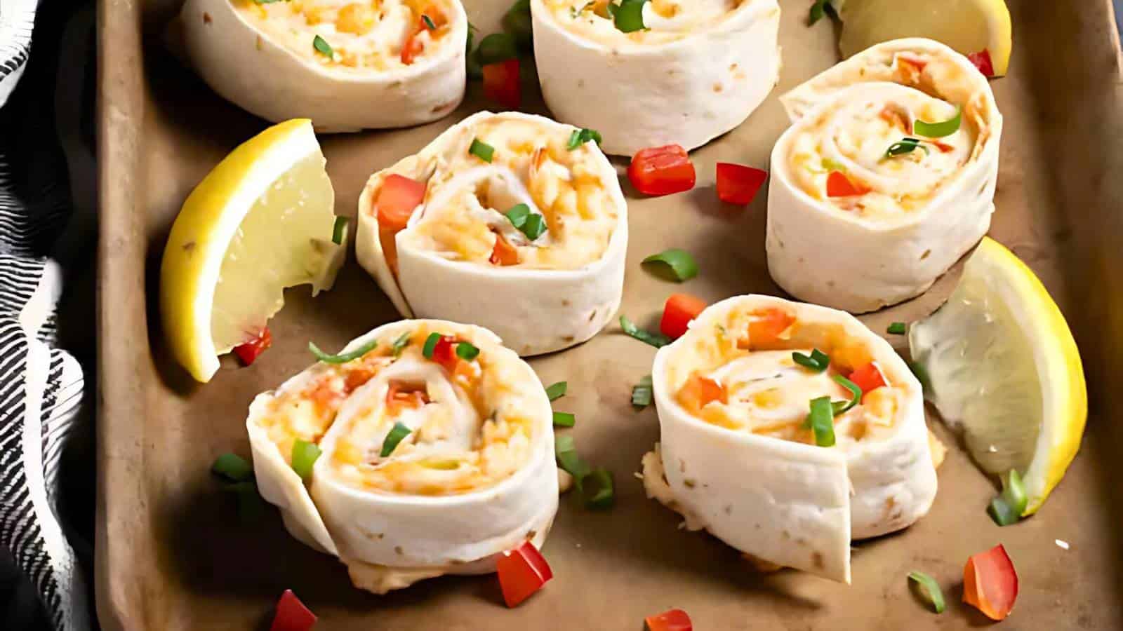 Six tortilla pinwheels filled with cheese and chopped vegetables are arranged on a baking tray, garnished with lemon wedges and diced red and green vegetables.