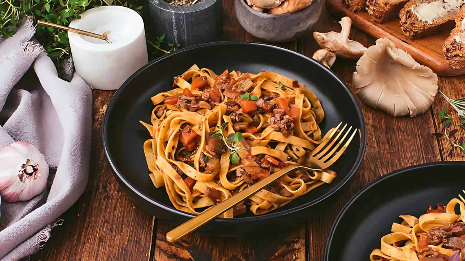 A black bowl of tagliatelle pasta with mushroom and vegetable sauce, garnished with herbs, placed on a wooden table with a gold fork and surrounding food items.