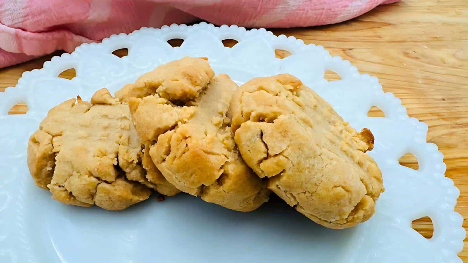 Three crumbly peanut butter cookies arranged on a white decorative plate, with a wooden surface and a pink cloth in the background.