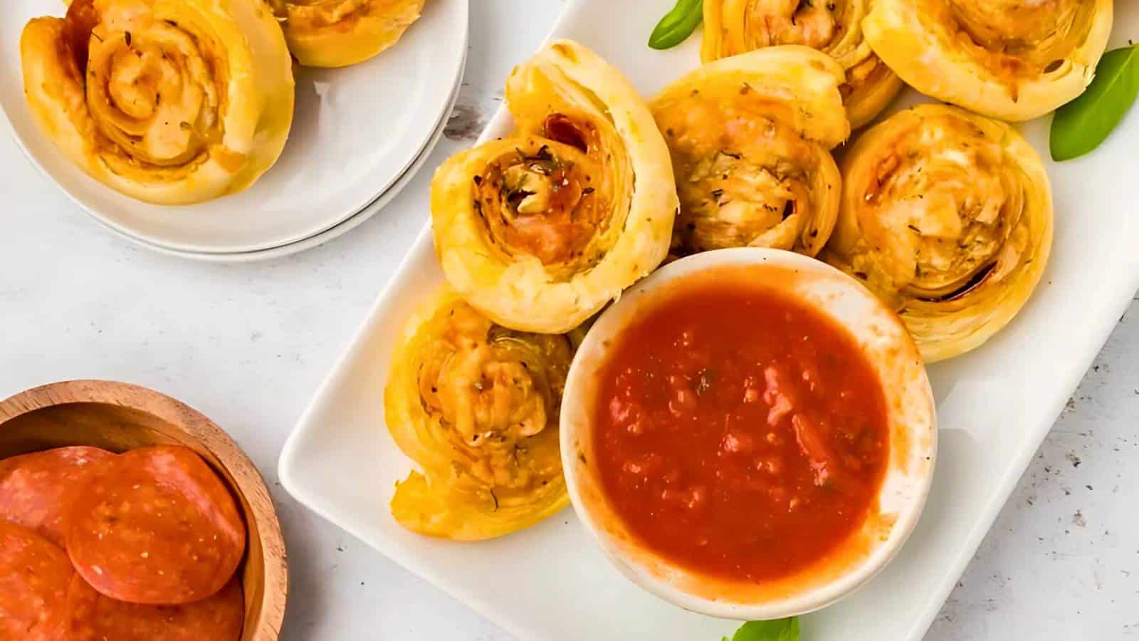 A white plate with pizza pinwheels arranged around a bowl of marinara sauce, with extra pinwheels and pepperoni slices on the side.