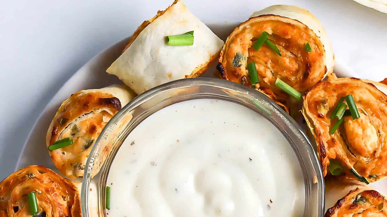 A plate with pinwheel-style appetizers filled with a reddish filling, garnished with chopped chives, arranged around a bowl of creamy white dipping sauce.