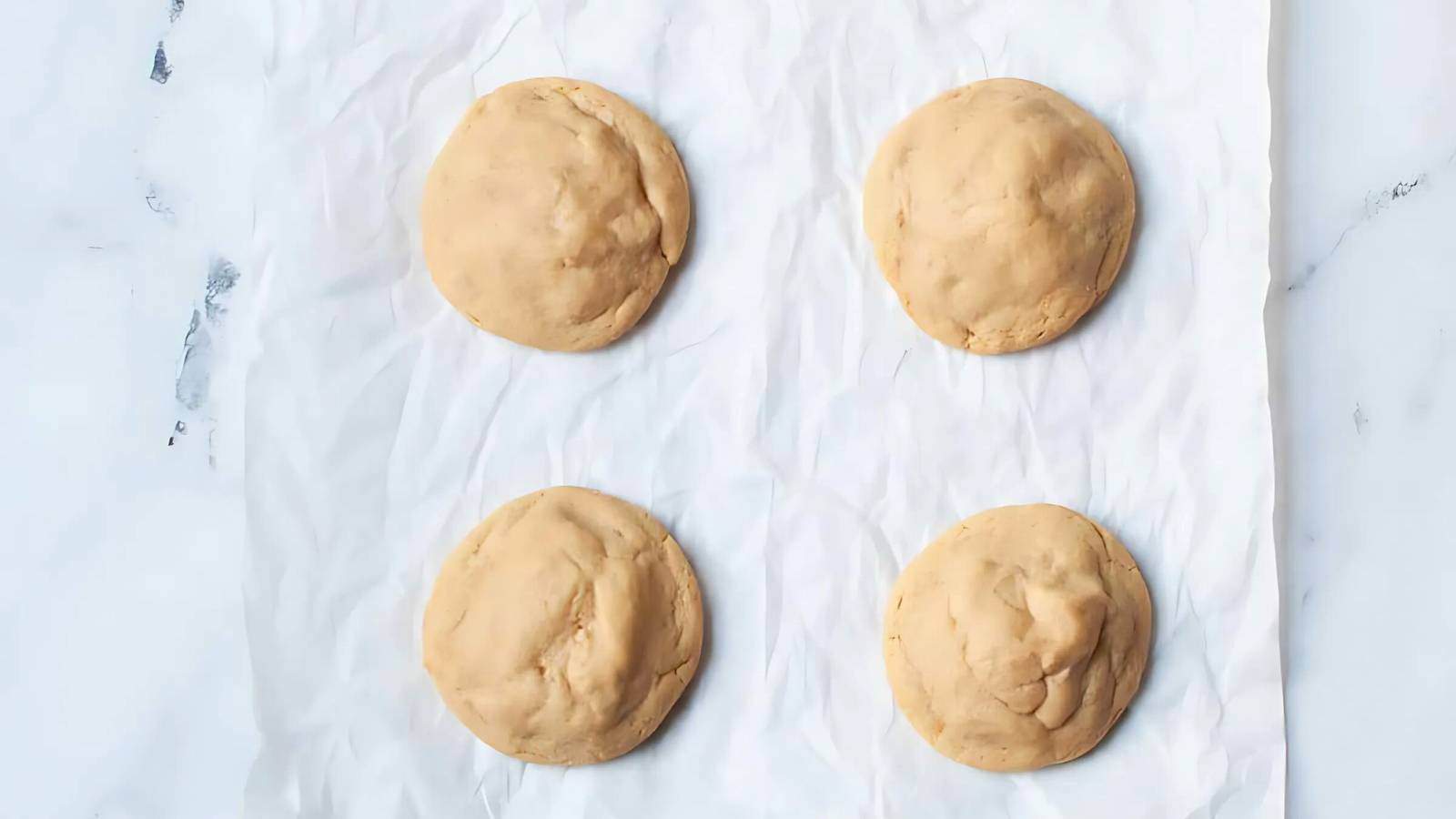 Four round, unbaked cookie dough balls are placed on a sheet of parchment paper set on a white surface.