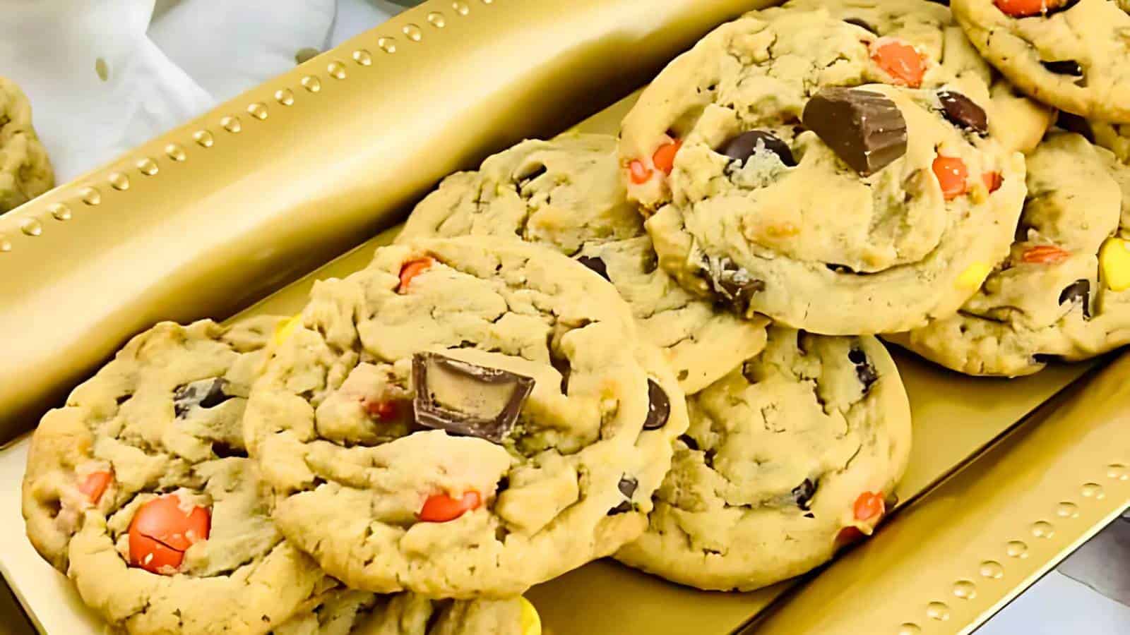 A close-up of a stack of large cookies with visible chocolate chunks and colorful candy pieces on a gold rectangular tray.