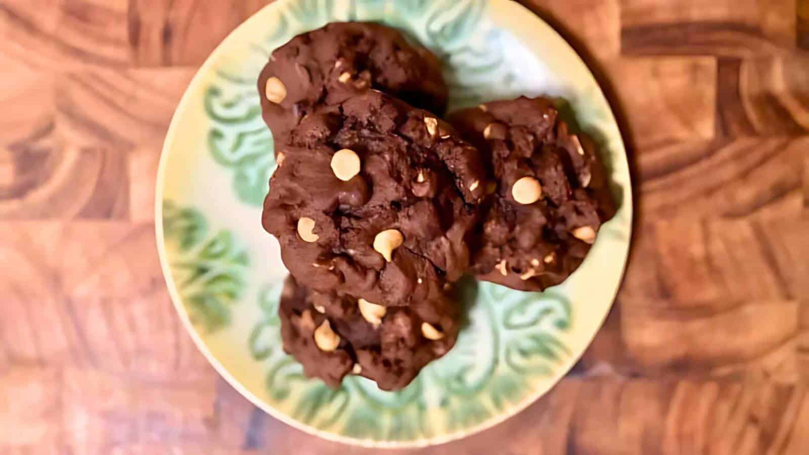 A plate with three chocolate cookies containing white chocolate chips, placed on a wooden surface.