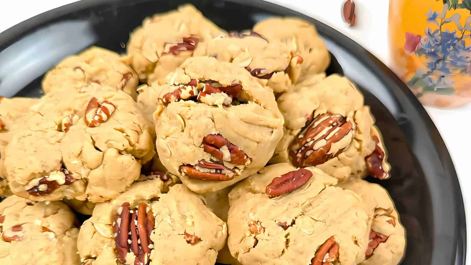 A black plate filled with pecan cookies, some with visible pecan halves on top, next to a glass with a floral design.