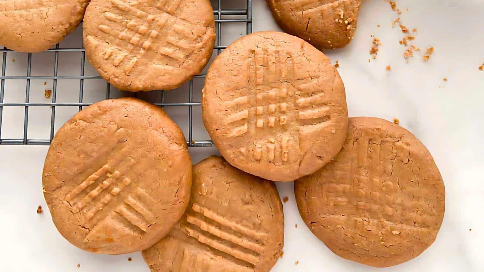 Seven round peanut butter cookies with fork marks are placed on a cooling rack and a white surface, with some crumbs visible.