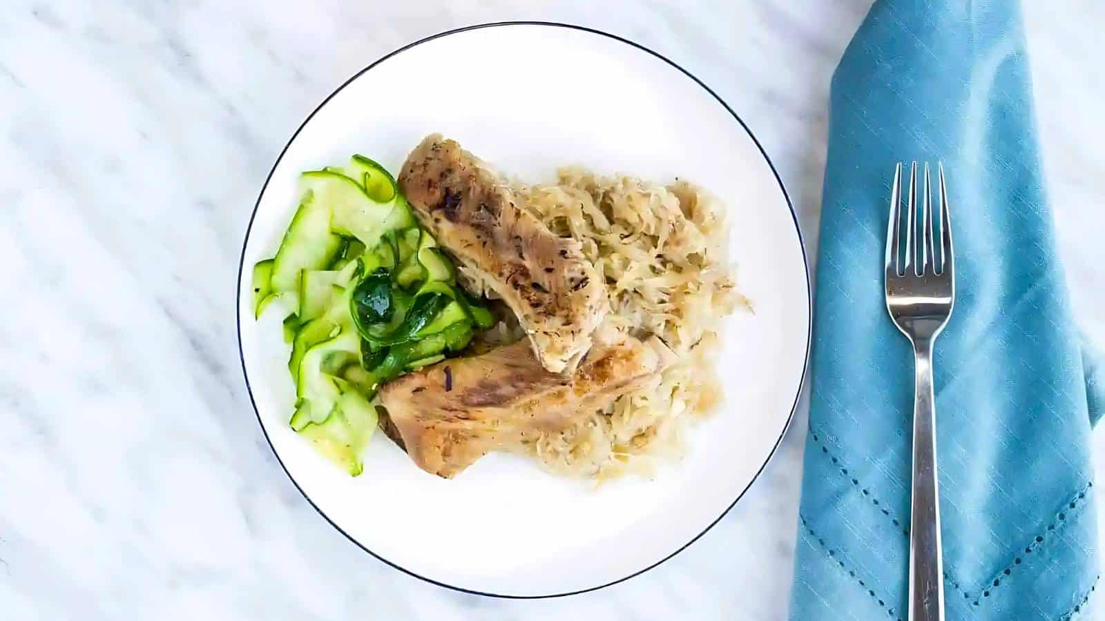 A white plate with two pieces of cooked meat, a portion of sauerkraut, sliced sautéed zucchini, and a blue napkin with a fork beside it.