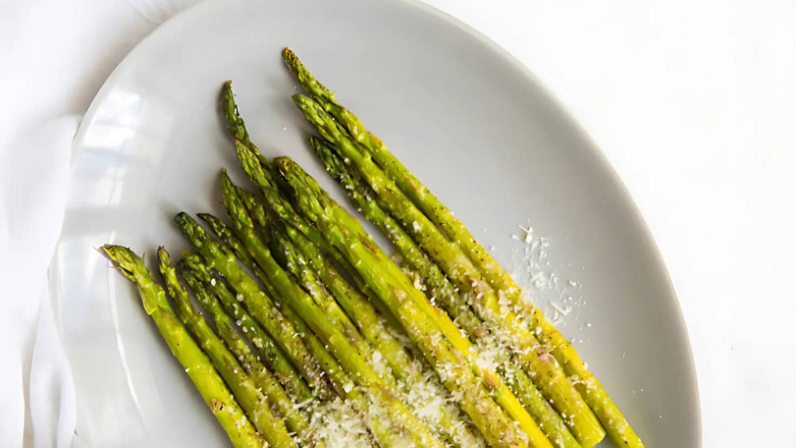 A plate of roasted asparagus spears topped with grated Parmesan cheese on a white background.
