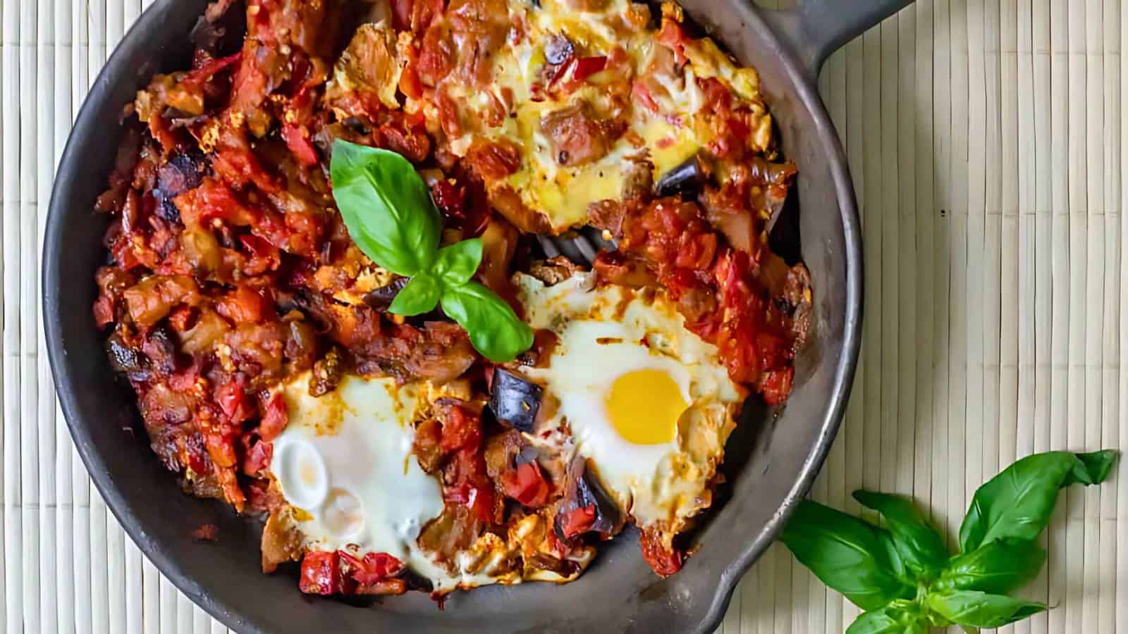 A skillet with baked eggs in a tomato and vegetable sauce, garnished with fresh basil leaves, is placed on a bamboo mat.