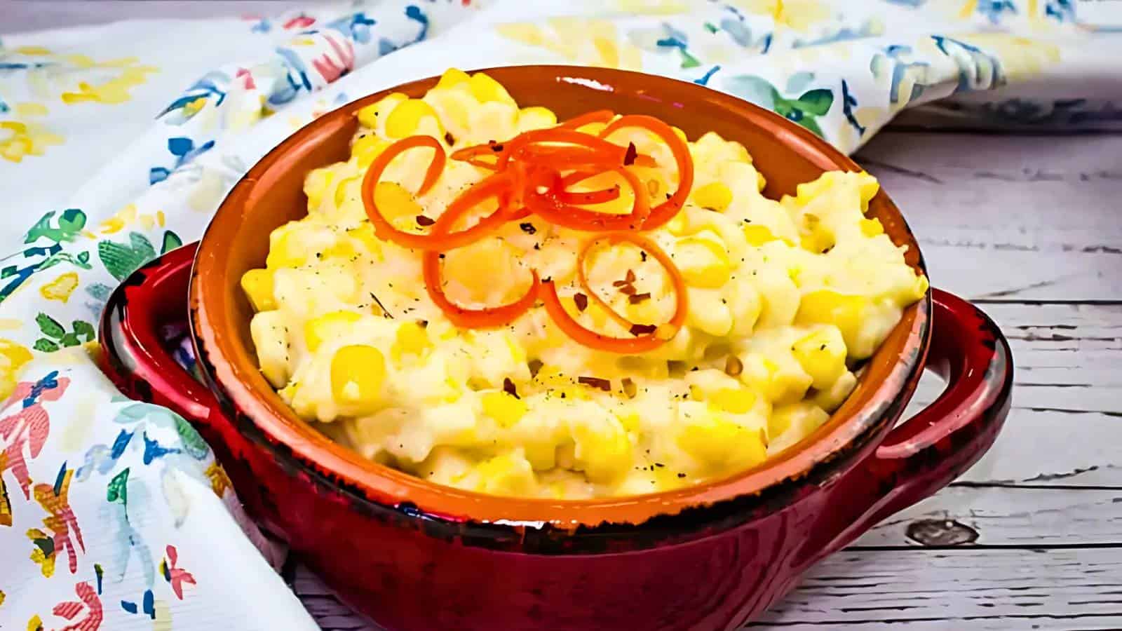 A red ceramic bowl filled with creamy corn pudding, topped with thin red pepper slices, sits on a wooden surface beside a patterned cloth.