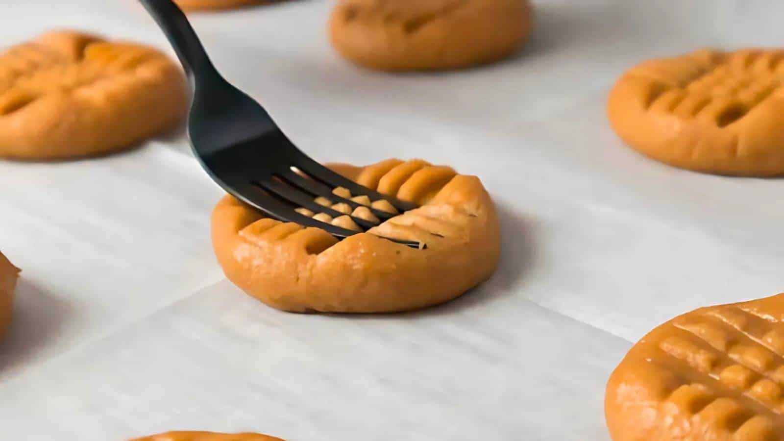 A fork is pressing a crisscross pattern into a round ball of cookie dough on a baking sheet lined with parchment paper.