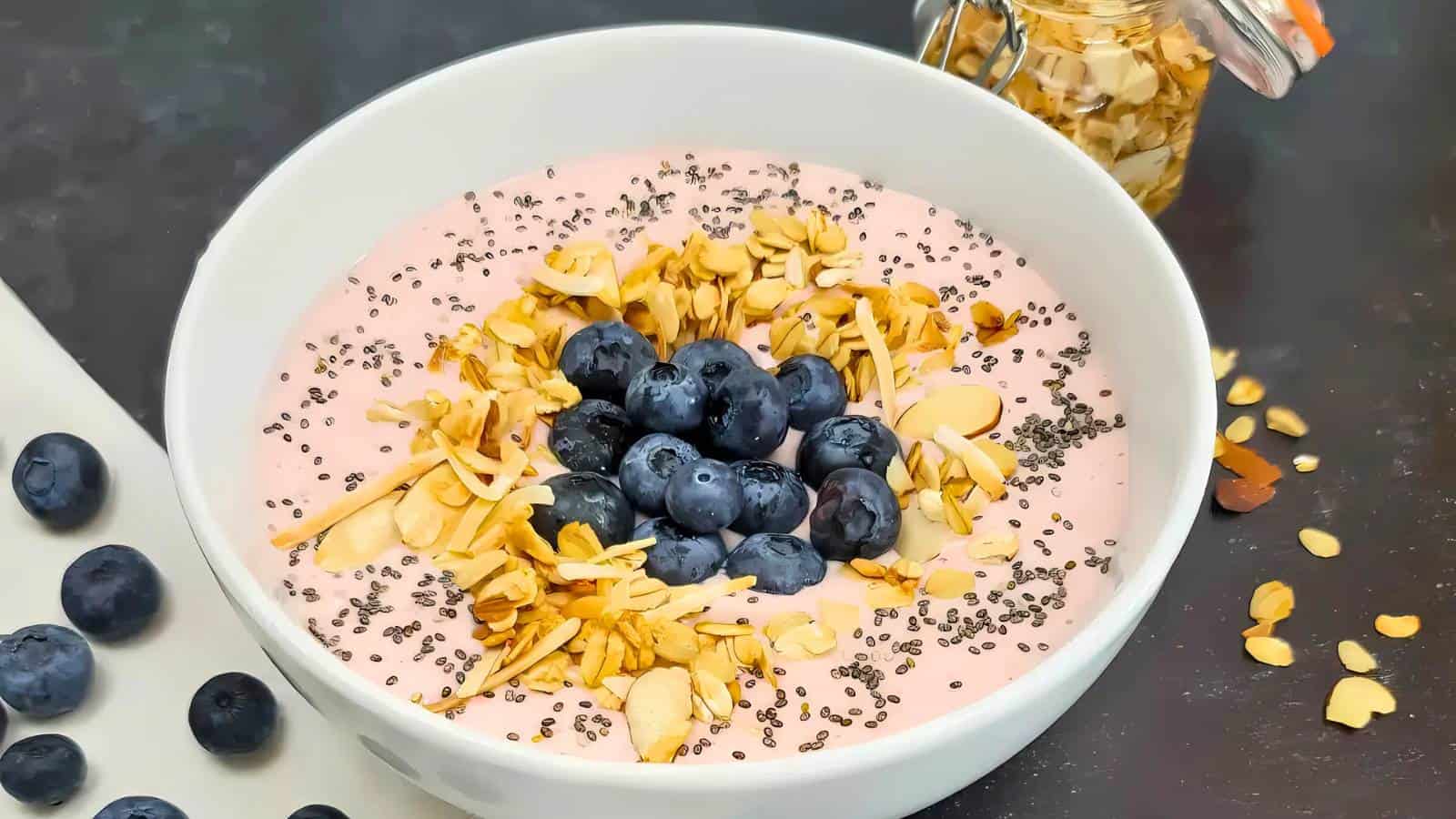 A bowl of pink smoothie topped with blueberries, sliced almonds, granola, and chia seeds on a dark surface, with loose blueberries and a jar of almonds nearby.
