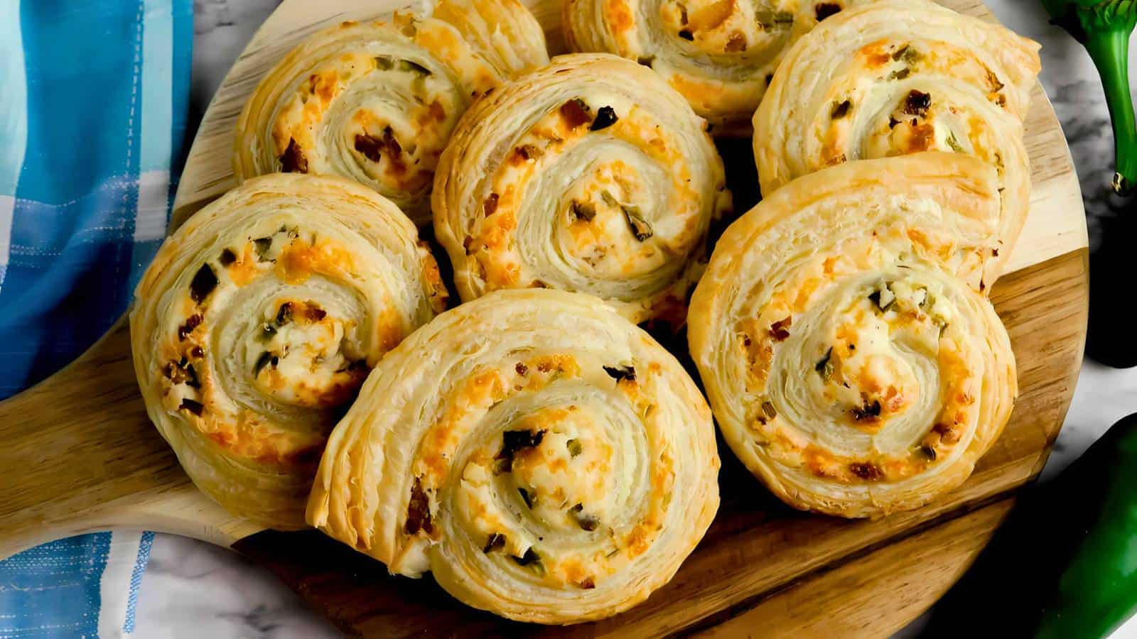 A wooden board with eight spiral-shaped puff pastries filled with cheese and herbs, displayed on a marble surface.