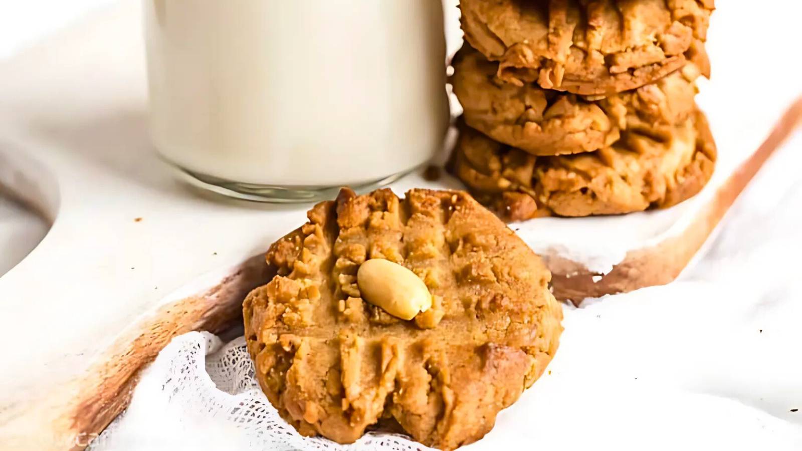 Peanut butter cookies, one topped with a peanut, rest on a wooden board near a glass of milk.